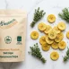 Packung salzige Kochbananenchips mit echten Alpenkräutern und Rosmarinzweigen auf Marmorhintergrund – veganer Premium-Snack aus Zürich.