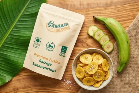 Kochbananenchips Alpensalz – Herzhafter Klassiker. Produkt Verpackung herzhafte Kochbananenchips Alpensalz auf rustikalem Holzgrund mit einer frischen grünen Kochbanane und grobem Salz – die knusprige Alternative zu Kartoffelchips.